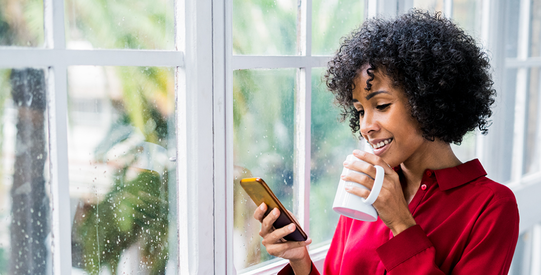 A woman holds a cup of coffee looking at her mobile phone screen happily